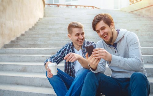 two men sitting down looking at their phone and smiling while drinking coffee