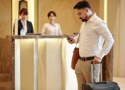 Serious elegant man with suitcase standing in hotel lobby and checking text messages in smartphone