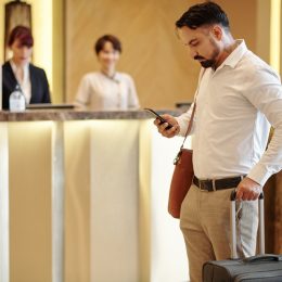 Serious elegant man with suitcase standing in hotel lobby and checking text messages in smartphone