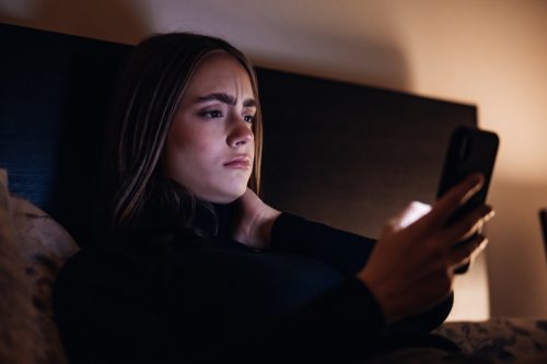 Worried and disappointed looking young woman lying on her bed in illuminated bedroom at night reading bad news in her e-mails, chat messages or social media posts on her Mobile Phone. Ambient Bedroom Night Lighting. Millenial Generation Modern Technology Lifestyle.