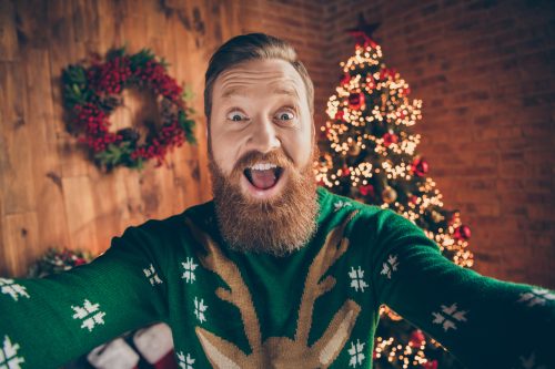 bearded man in christmas sweater taking selfie in front of christmas tree. 