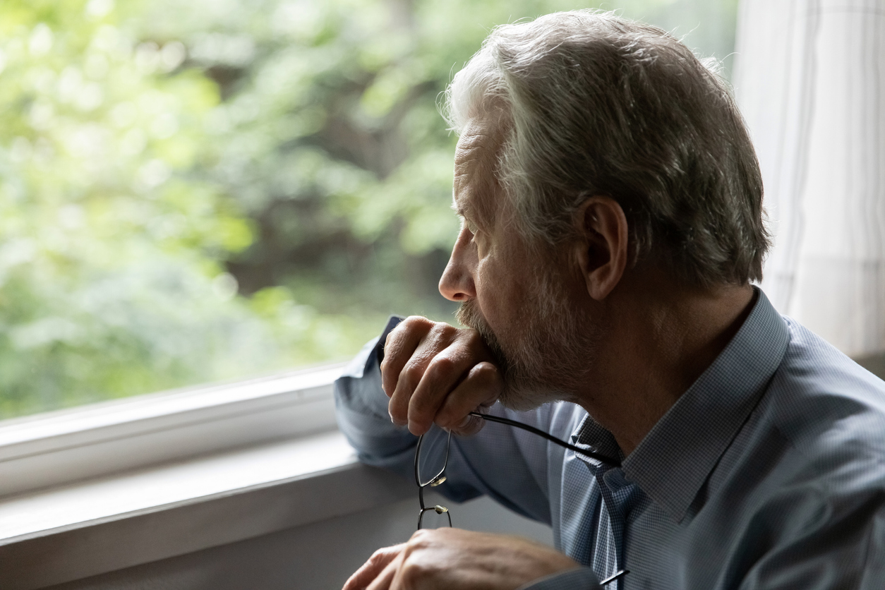 Man in deep thoughts holds glasses, looks out window. 