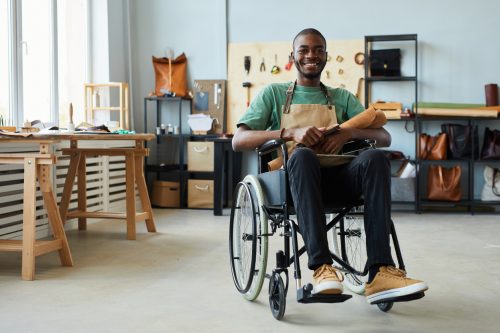 Full length portrait of smiling African-American man using wheelchair enjoying work in leatherworkers workshop and looking at camera