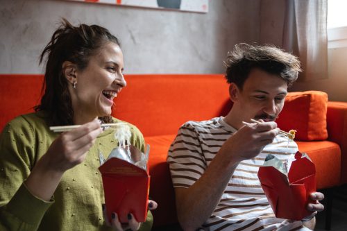 Modern young Caucasian couple enjoy the Chinese take out food, while sitting on the floor and having fun