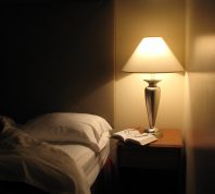 Bed next to night table with a lamp turned on.