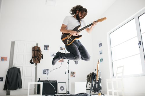 One man, playing guitar and jumping, in home studio.