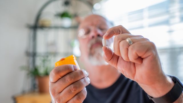 Mature Man Scrutinizing His Perscription Medications Holding a Pill in One Hand and the Bottle in the Other In a Modern Home
