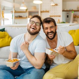 A male couple eating pasta out of takeout containers while sitting on their living room floor and laughing.