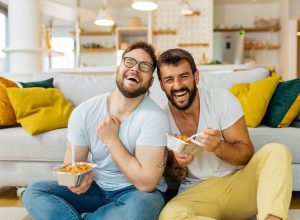 A male couple eating pasta out of takeout containers while sitting on their living room floor and laughing.