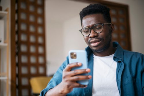 Young man sitting at home, feeling depressed and trying to contemplate bad news he is reading online using a smart phone