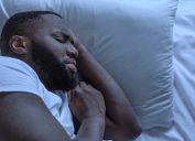 Close up of a man in a white t-shirt sleeping on white sheets looking anxious.