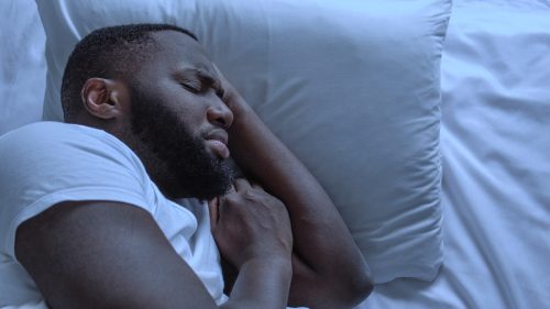 Close up of a man in a white t-shirt sleeping on white sheets looking anxious.