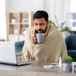 sick young man wrapped in blanket with laptop computer drinking hot tea and working at home