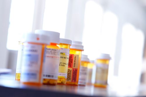 Several prescription medication bottles sit on a table. Light pours in through the windows in the background bathing the room with a soft glow. The image is photographed with a very shallow depth of field.