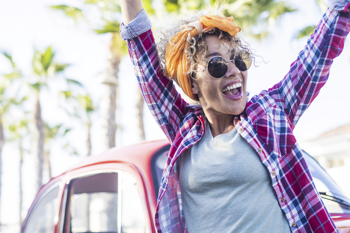 A happy young woman smiling and throwing her arms up in front of a red car.