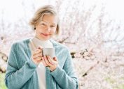 An elegant older woman holding a mug and standing against a background of cherry blossom trees.