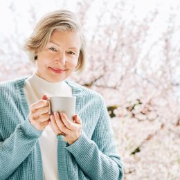 An elegant older woman holding a mug and standing against a background of cherry blossom trees.