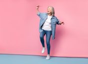 An older, gray-haired woman wearing skinny jeans and a denim shirt dancing happily on a pink background.