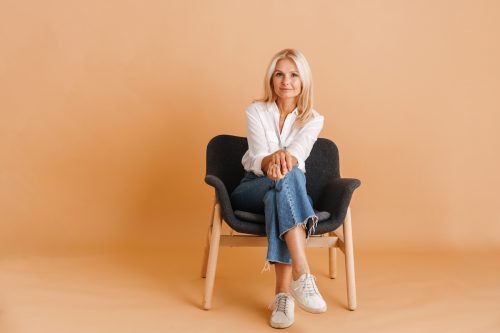 An older blonde woman wearing jeans and a white shirt sitting on a chair against an orange background.