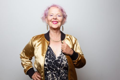 An eccentrically dressed older woman with pink hair wearing a leopard-print dress and gold jacket