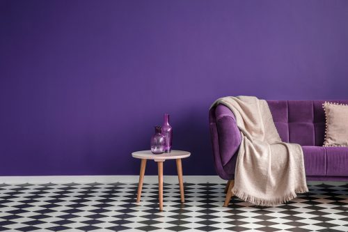 Dark purple sofa with a blanket beside a small table with bottles standing on black and white checkerboard floor in a minimalistic living room with purple walls.