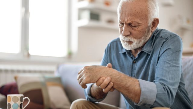 A senior man sitting on the couch and rubbing his hand due to arthritis.