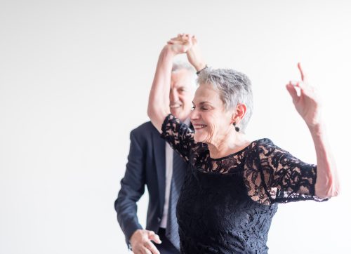 Close-up of an elegantly dressed older man and woman dancing exuberantly against a neutral background