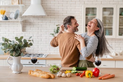 Happy cheerful middle-aged mature couple dancing together in the kitchen, preparing cooking food meal for romantic dinner, spending time together.
