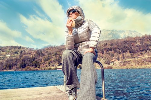 mature woman in puffer coat eating a sandwich on a lake