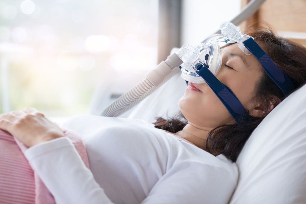 Senior woman using cpap machine to stop choking and snoring from obstructive sleep apnea with bokeh and morning light background. Woman and cpap mask, healthcare concept.