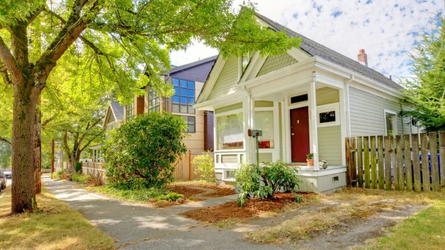 A cute green and white craftsman house with a red door, located on a tree-lined street.
