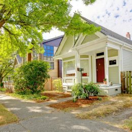A cute green and white craftsman house with a red door, located on a tree-lined street.