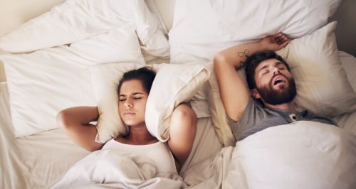 Shot of a young woman covering her ears with a pillow while her husband snores in bed