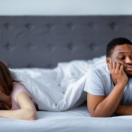 A young couple under the covers at the foot of the bed, each looking away from each other and upset.