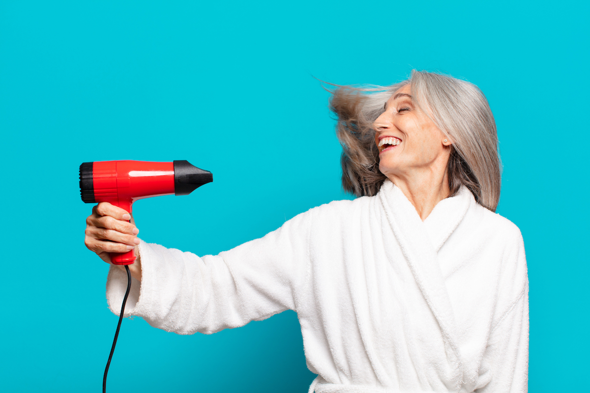 An elderly woman with gray hair in a white coat uses a blow dryer against a teal background.