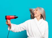 An older woman with gray hair in a white robe using a blow dryer against a teal background.