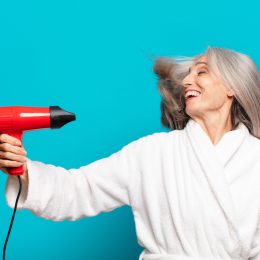 An older woman with gray hair in a white robe using a blow dryer against a teal background.