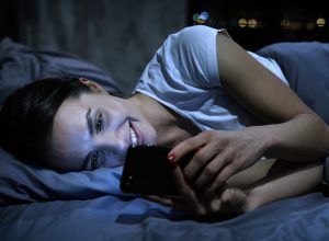 A young female smiling while on her phone at night in bed.