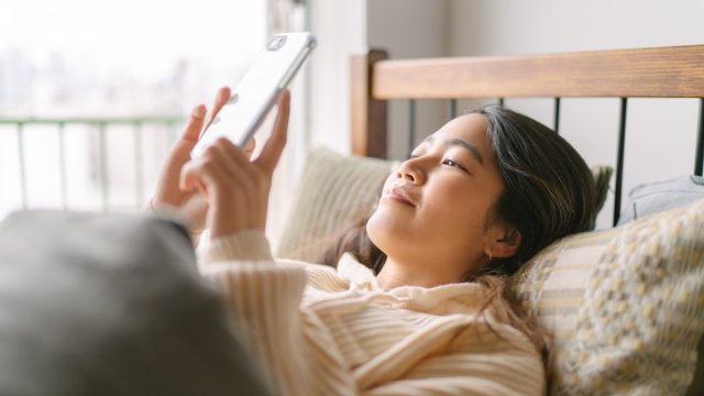 A young and beautiful woman is lying in bed and using a smart phone happily in her bed in the morning.