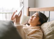 A young and beautiful woman is lying in bed and using a smart phone happily in her bed in the morning.