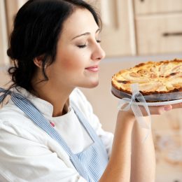 A young woman in an apron smells a freshly baked apple pie.