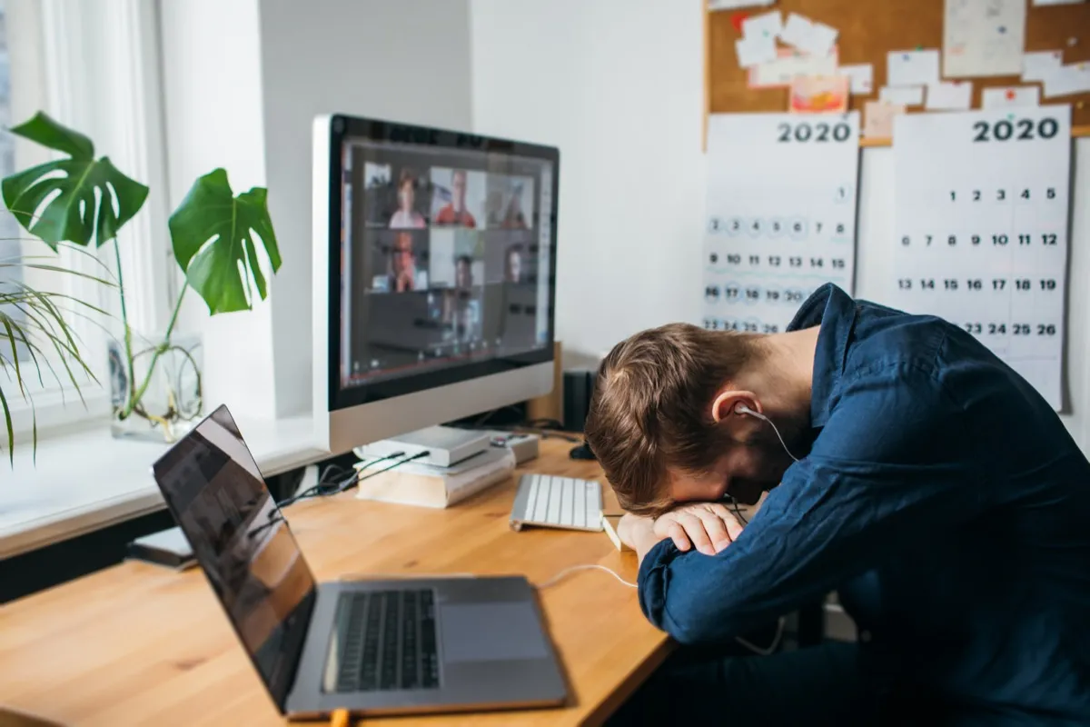 man sleeping with his head on his deck while a zoom call is up on his computer