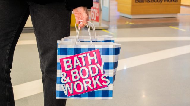 A female customer holds in her hand paper bags branded "Bath and body works" in a blue and white check and a pink logo on the background of the entrance to the store