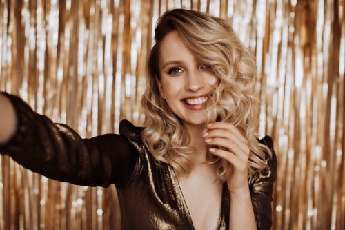 A smiling blonde woman wearing a bronze metallic dress against a gold party background.