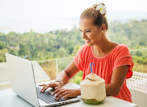 A young blonde woman working on her laptop with a tropical background and a coconut sitting next to her.