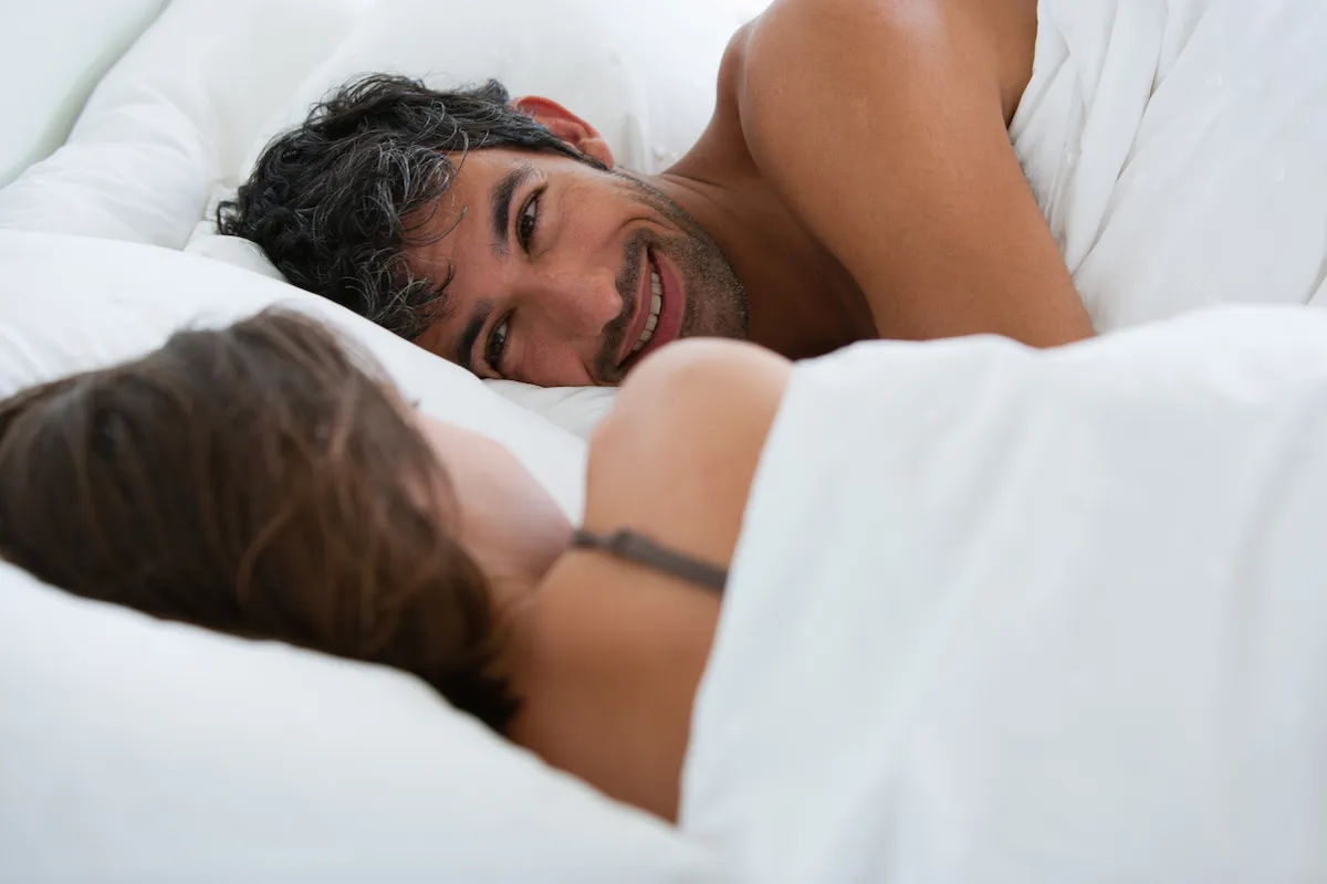 A couple looking at each in bed, with the man's face shown smiling.