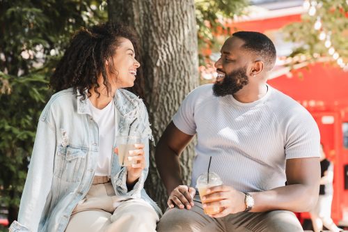 A couple on a date is sitting outside in front of a tree smiling while drinking lemonade.