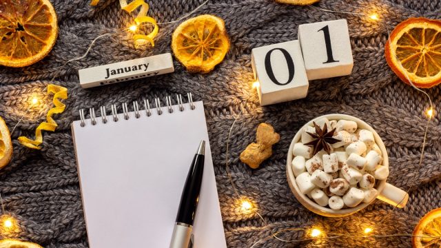 Winter composition. Wooden calendar January 1st Cup of cocoa with marshmallow, empty open notepad with pen, dried oranges, light garland on grey knitted background. Top view Flat lay Mockup