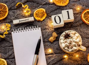 Winter composition. Wooden calendar January 1st Cup of cocoa with marshmallow, empty open notepad with pen, dried oranges, light garland on grey knitted background. Top view Flat lay Mockup