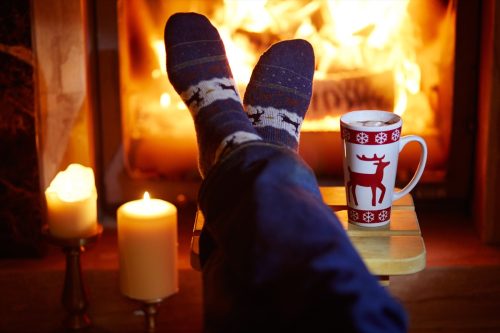 Man's feet in warm socks with large mug of hot chocolate and murshmallows near fireplace. Cozy Christmas evening